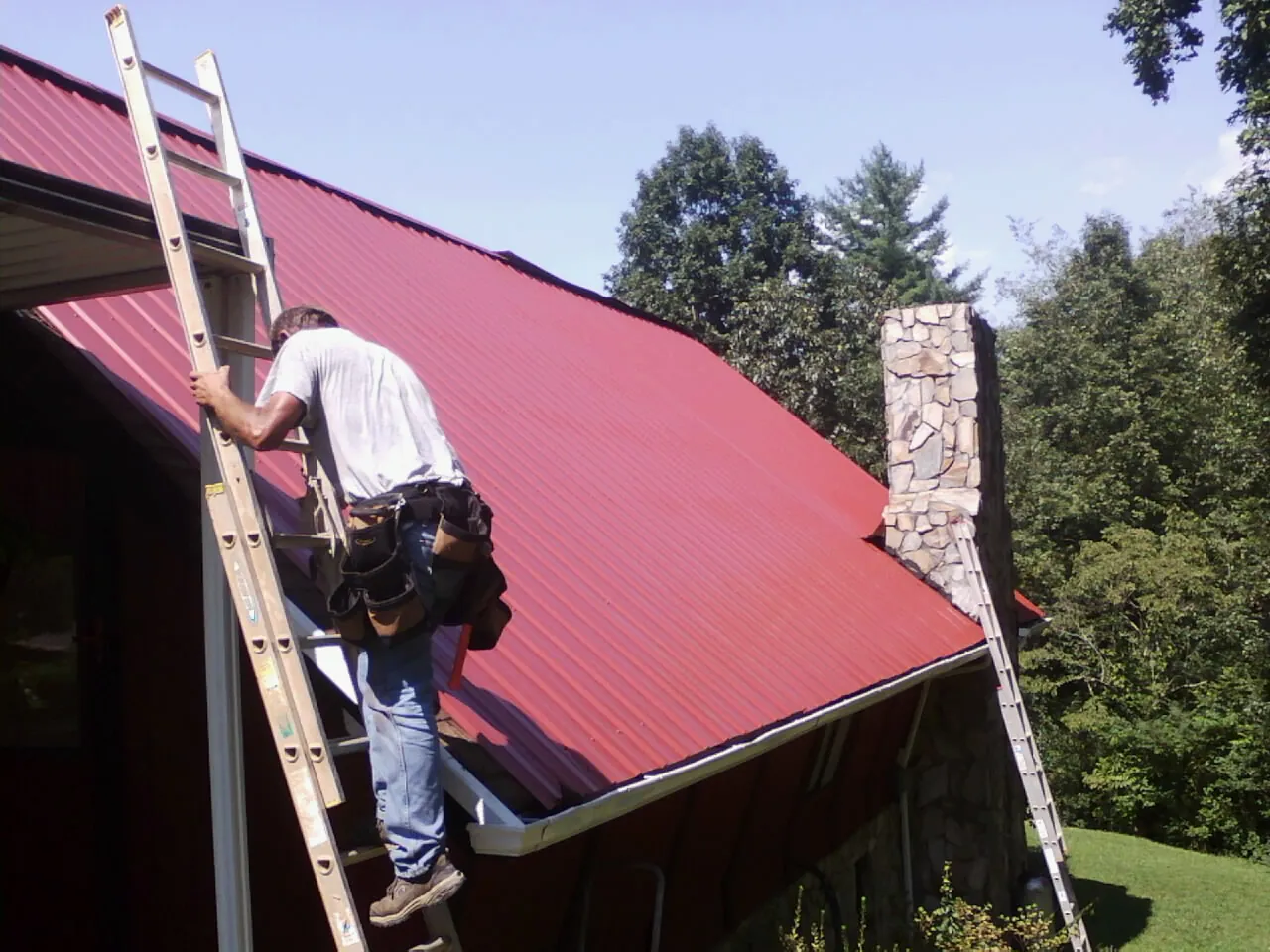 Roofer on a ladder beside a red metal roof and stone chimney — Soffit & Fascia Repair by Roofing Brothers in Woosung